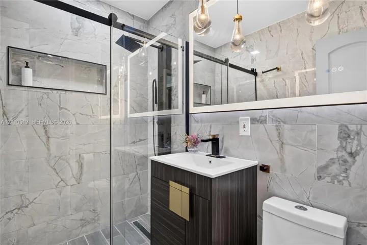 Bathroom featuring full-height large-format stone tile, a glass-enclosed shower with a built-in recessed wall niche, a dark wood-finish vanity with a rectangular sink, matte black fixtures, and a framed mirror with pendant lighting