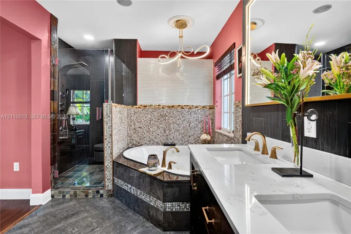 Bathroom featuring a dual-sink vanity with white stone-finish countertops, gold-toned fixtures, a corner jetted soaking tub with mosaic tile surround, and an enclosed shower with dark tile walls