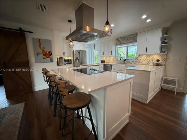 Kitchen featuring white shaker-style cabinetry, an expansive central island with a solid-surface countertop and built-in cooktop, stainless steel vent hood, apron-front sink, and wood-finish flooring