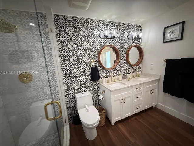 Bathroom featuring a dual-sink vanity with light-toned stone countertops, brass-finish fixtures, dual round mirrors, wood-finish flooring, and a patterned tile feature wall