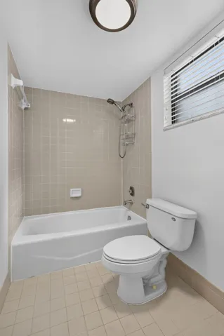 Bathroom featuring a built-in bathtub with a tile surround, window with horizontal blinds, flush-mount lighting, and neutral floor tiling