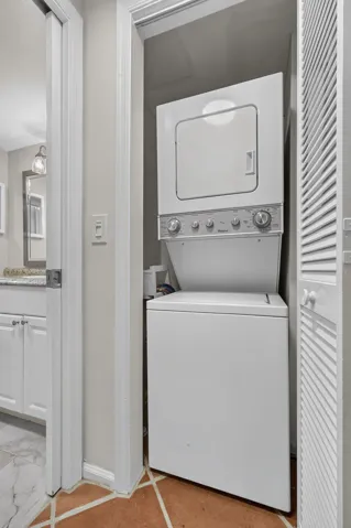 Stacked laundry unit featuring white appliances housed within a closet with a louvered door, complemented by earth-toned tile flooring