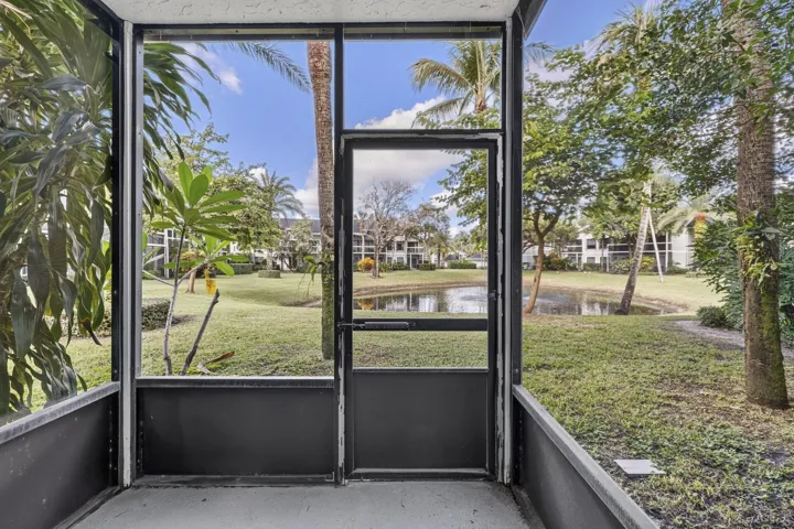 Screened-in enclosure featuring a metal-frame door, concrete flooring, and direct access to a grassy common area with a pond and mature palm trees