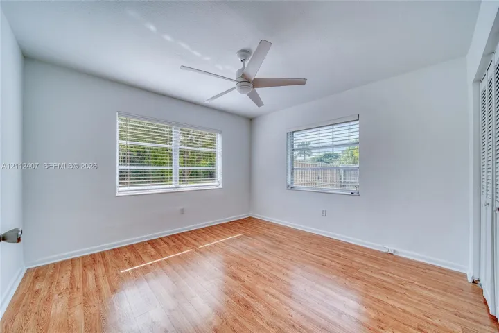 Neutral-toned room featuring wood-finish flooring, dual windows with horizontal blinds, a ceiling fan, and louvered closet doors