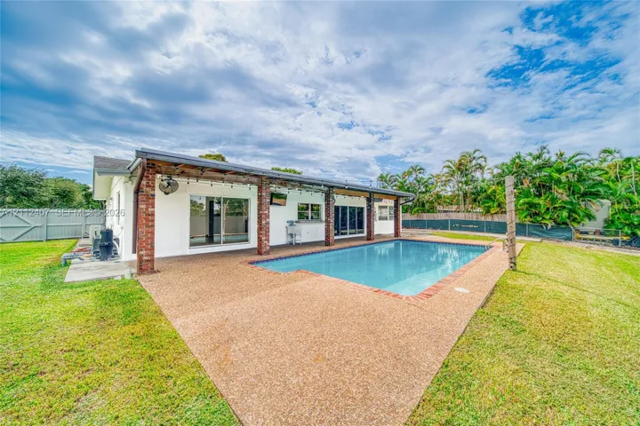 Rear exterior featuring a covered patio with brick support columns, an in-ground pool with brick coping, expansive lawn, and fenced perimeter