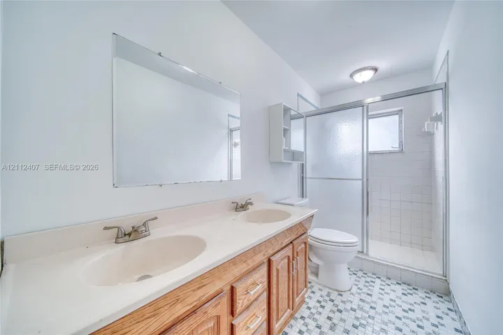 Dual-sink vanity with a molded countertop, wood-finish cabinetry, frameless wall-mounted mirror, and walk-in shower with a glass enclosure