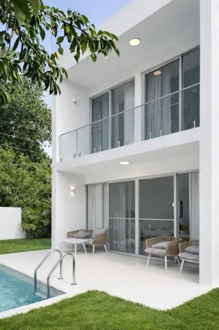 Contemporary two-story exterior featuring white stucco finish, second-level glass-railing balcony, recessed lighting, and integrated sliding glass doors leading to a paved patio with an in-ground pool