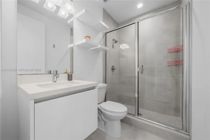 Bathroom featuring a white vanity with a rectangular sink, quartz countertop, built-in wall shelving, and a tiled shower enclosure with a glass door