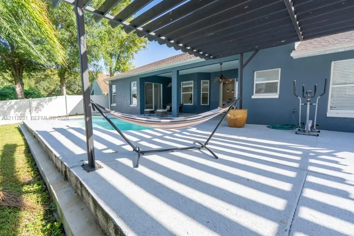 Covered concrete patio featuring a dark-toned pergola, built-in swimming pool, and white perimeter privacy fencing