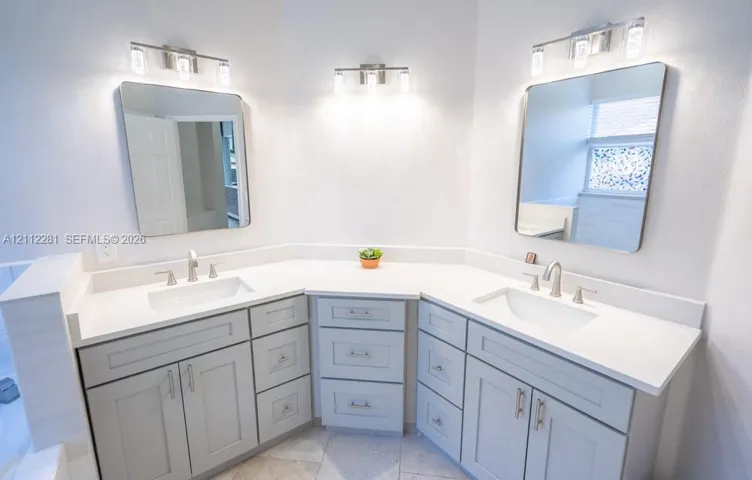 Double vanity featuring grey shaker-style cabinetry, white solid-surface countertops, integrated rectangular sinks, brushed nickel faucets, and two three-light vanity fixtures