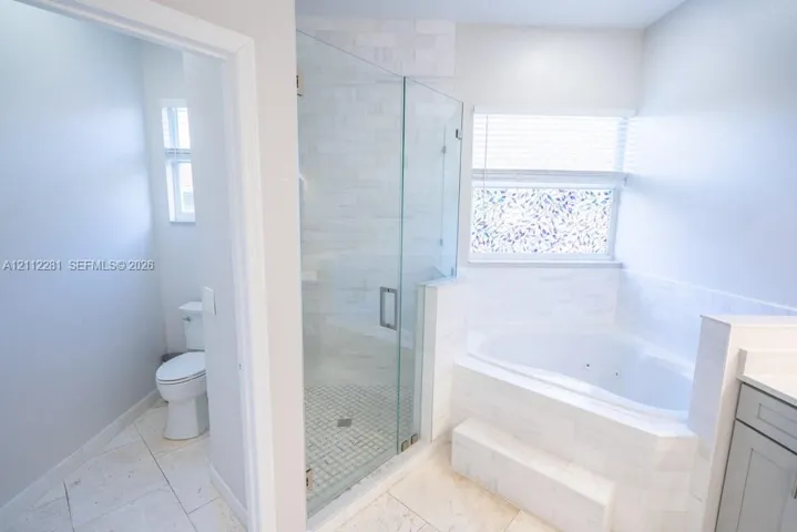 Walk-in glass-enclosed shower featuring stone-tile surround, oversized soaking tub with tile deck, light-finish tile flooring, and integrated commode area