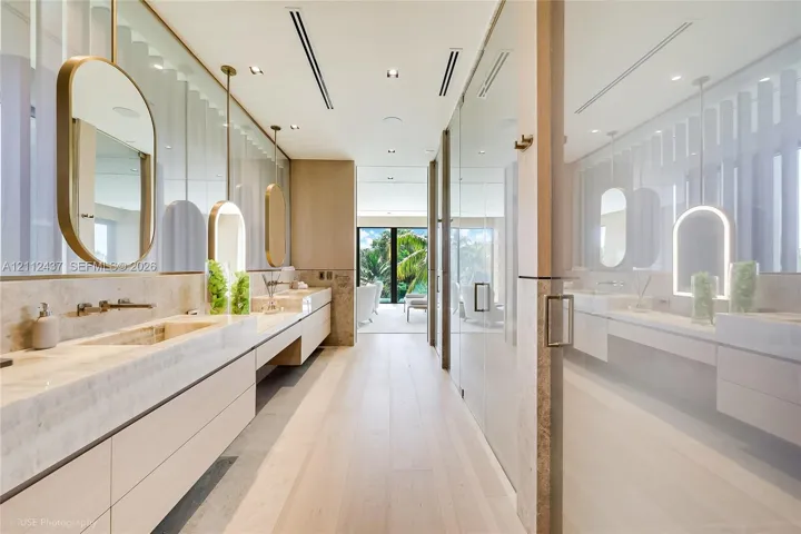 Dual floating vanities with integrated stone sinks and wall-mounted faucets, suspended oval mirrors, recessed ceiling lighting, wood-finish flooring, and floor-to-ceiling glass paneling