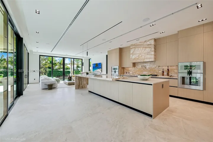 Custom wood-finish cabinetry, expansive stone-topped kitchen island with waterfall edge, integrated stainless steel appliances, and floor-to-ceiling glass sliding doors