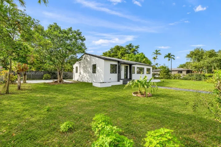 Single-story residence featuring a smooth white stucco exterior, dark-toned window trim, an expansive grass lawn, and mature canopy trees
