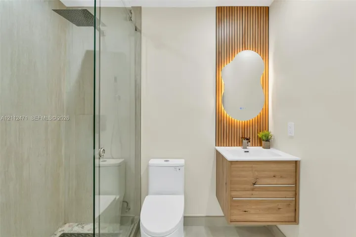 Walk-in glass enclosed shower with rainfall showerhead, floating wood-finish vanity, integrated solid surface sink, and decorative vertical wood-slat wall feature with backlit organic-shaped mirror