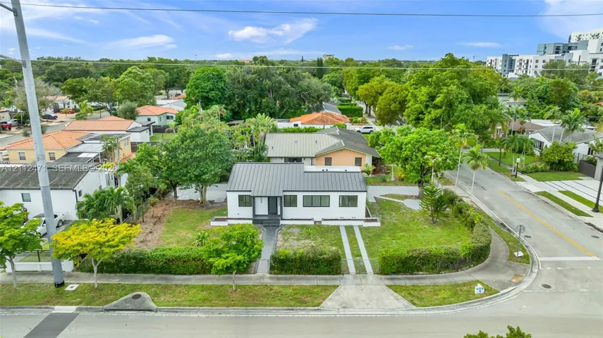 Single-story residence featuring a standing-seam metal roof, white stucco exterior, dark-trimmed windows, a paved walkway, and a landscaped corner lot