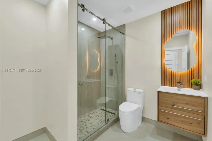Glass-enclosed shower with a pebble-stone floor, wood-finish floating vanity, vertical wood-slat accent wall, and irregular-shaped backlit mirror