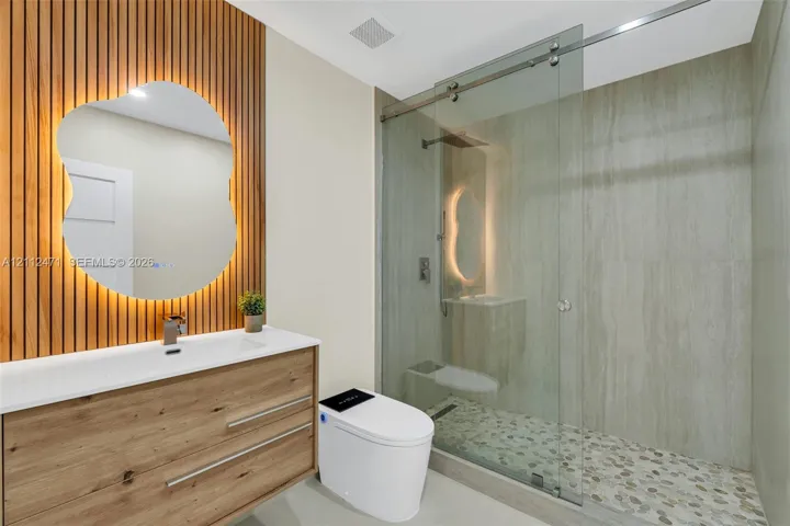 Wood-slat accent wall featuring a backlit, organic-shaped mirror, wood-finish vanity with an integrated basin, and a walk-in shower with a glass enclosure and pebble-stone flooring