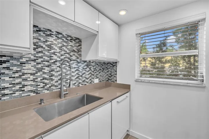 Glossy white cabinetry, solid-surface countertop, stainless steel undermount sink with high-arc faucet, and multi-tone glass tile backsplash