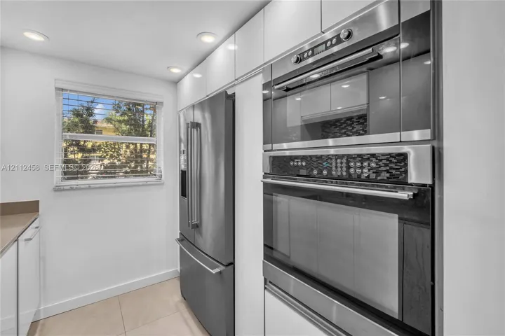 Kitchen featuring high-gloss white cabinetry, integrated stainless steel appliances, a built-in wall oven and microwave unit, recessed ceiling lighting, and large-format tile flooring