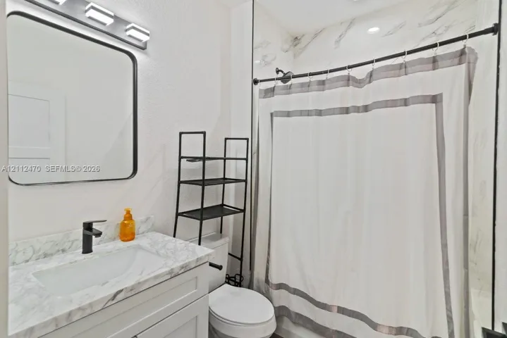 Bathroom vanity with stone-finish countertop, undermount sink, matte black faucet, rectangular mirror with a black frame, and shower surround with stone-finish wall tile