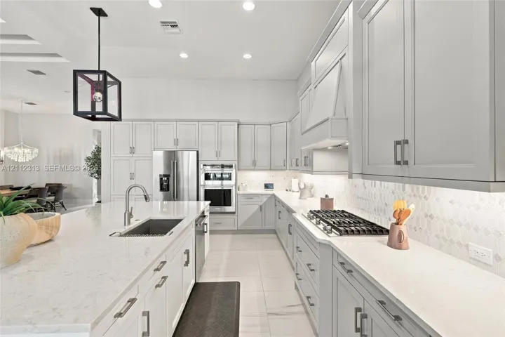 Kitchen featuring light-toned cabinetry, a spacious island with a built-in sink, stone-finish countertops, stainless steel appliances, and a geometric pendant light fixture