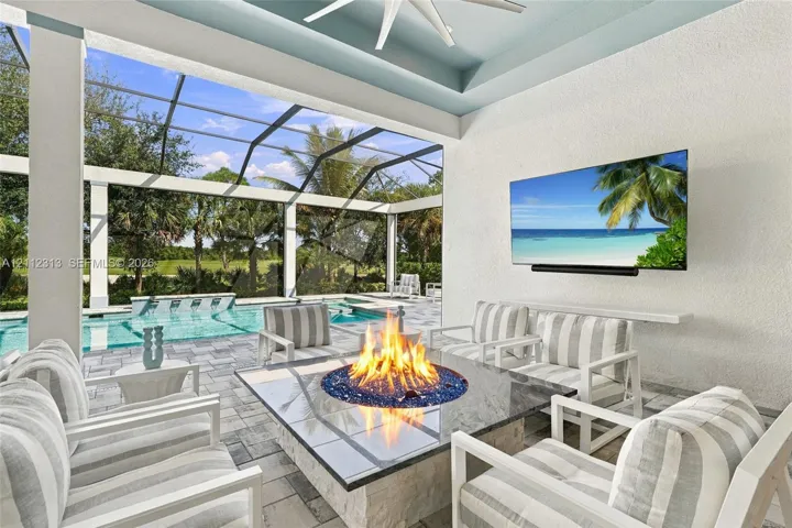 Covered lanai featuring a stone-topped built-in fire pit, screened enclosure, custom ceiling fan, and built-in wall-mounted television, accented by paver flooring and an integrated swimming pool