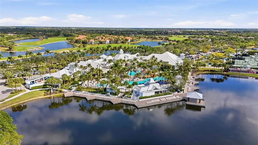 Aerial view of a coastal-inspired complex featuring a resort-style swimming pool, expansive waterfront sun deck, multi-tiered white roofing, and manicured tropical landscaping