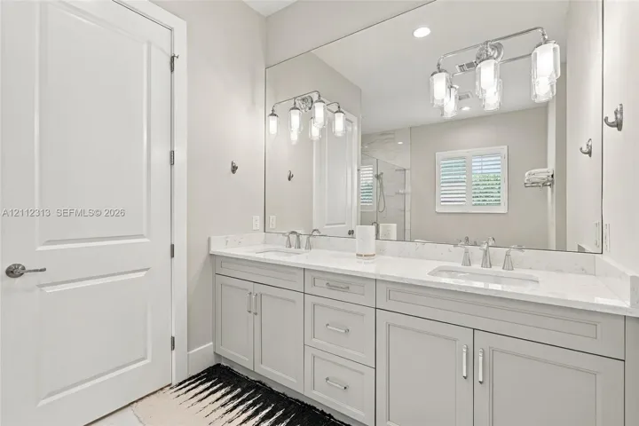 Double vanity featuring light-toned cabinetry, stone-finish countertops, undermount sinks, polished metal fixtures, and a wall-to-wall mirror with multi-bulb integrated lighting