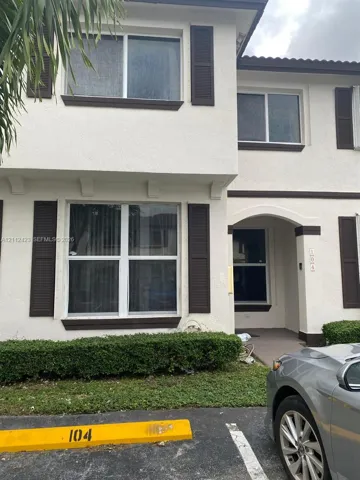 Two-story exterior featuring a light-toned stucco facade, dark decorative window shutters, an arched entryway, and manicured foundation shrubbery