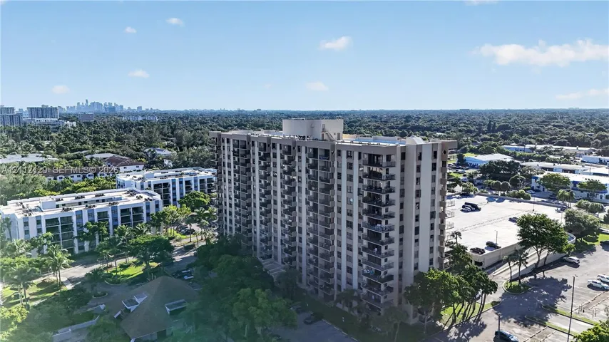 High-rise building featuring private balconies, expansive paved parking, mature landscaping, and an exterior swimming pool