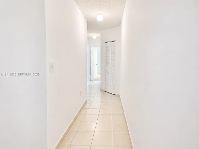 Hallway featuring neutral-toned tile flooring, white walls, and a bi-fold louvered closet door with flush-mount ceiling lighting