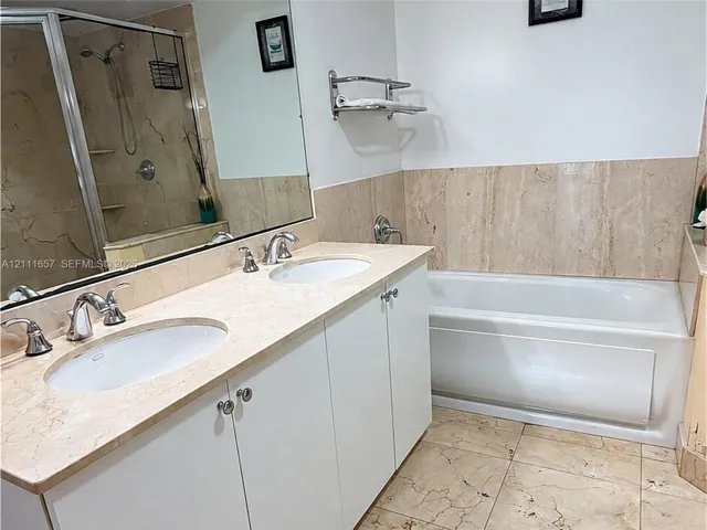 Dual-sink vanity with stone countertop, drop-in soaking tub with stone surround, tile flooring, wall-to-wall mirror, and glass-enclosed shower