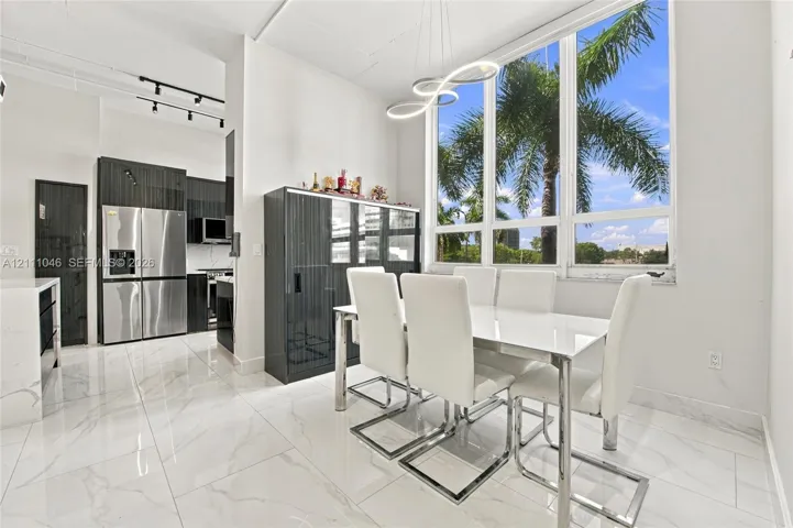 Open-concept area featuring floor-to-ceiling windows, white stone-finish tile flooring, custom dark-toned cabinetry, stainless steel refrigerator, and modern geometric pendant lighting