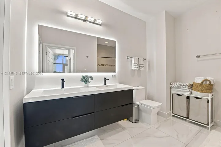 Floating double vanity with integrated sinks, matte black fixtures, illuminated mirror, and polished stone-finish floor tiling