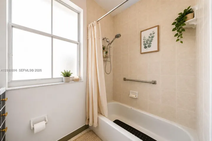 Full bathroom featuring a white porcelain tub, wall-mounted brushed metal grab bar, adjustable shower head, and neutral tiled surround