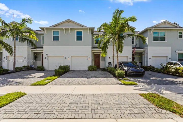 Multi-level exterior featuring light-toned siding, attached garages, stone-paver driveways, and mature palm trees