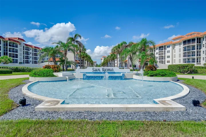 Gated community entrance featuring a tiered water fountain, mosaic tile accents, mature palm trees, and manicured landscaping