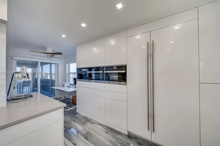 Sleek, high-gloss white cabinetry featuring integrated, full-height refrigerator and flush-mount wall ovens, quartz countertops, recessed ceiling lighting, and polished stone-finish flooring