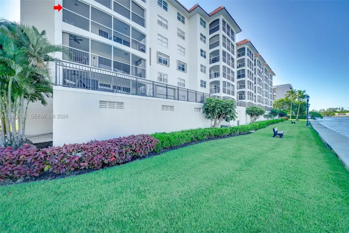 Multi-story building exterior featuring screened-in balconies, a stucco facade, red tile roofing, and direct waterfront grounds with manicured landscaping