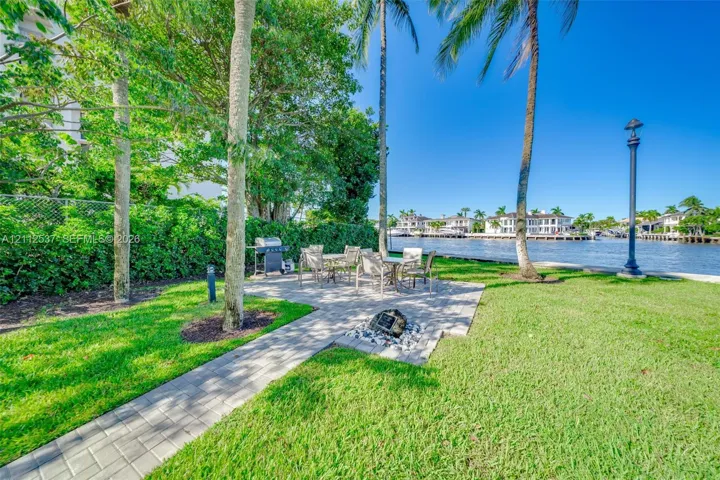 Waterfront grounds featuring a paver patio, manicured lawn, mature palm trees, and a decorative stone-lined fire pit area