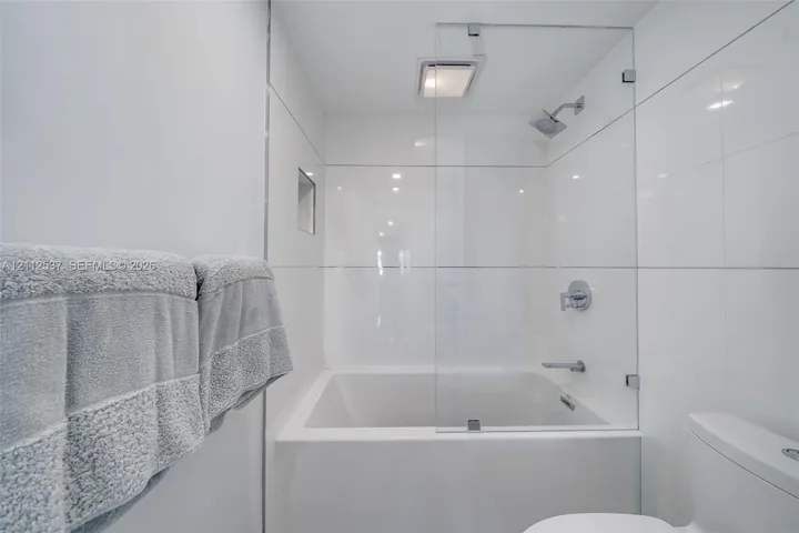 Soaking tub with a frameless glass splash panel, chrome rainfall showerhead, recessed wall niche, and floor-to-ceiling white tile surround