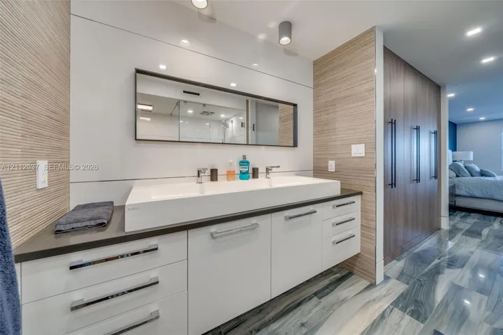 Modern floating vanity featuring a wide rectangular double-basin sink, high-gloss white cabinetry, integrated drawer pulls, wood-finish wall paneling, and recessed ceiling lighting