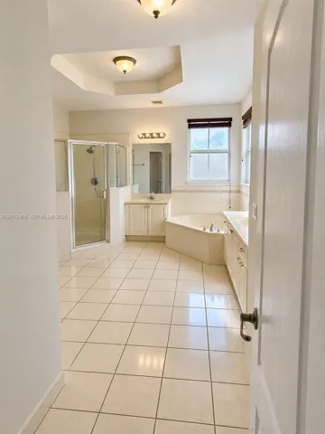 Ceramic tile flooring, corner soaking tub with tile surround, glass-enclosed shower, dual-sink vanity with light-colored cabinetry, and recessed tray ceiling