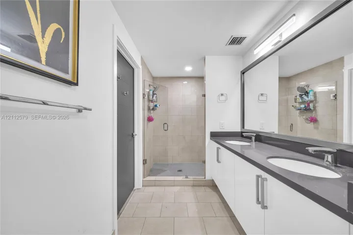 Dual-sink vanity with high-gloss cabinetry and stone-finish countertop, frameless glass shower enclosure with neutral-toned wall tile, and full-width vanity mirror with integrated lighting