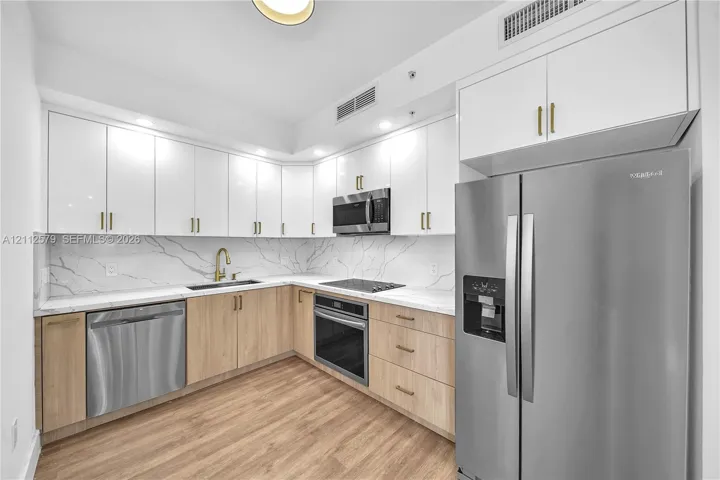 Kitchen featuring high-gloss white upper cabinetry, wood-finish base cabinetry, veined stone-look backsplash and countertops, brushed brass hardware, and stainless steel appliances