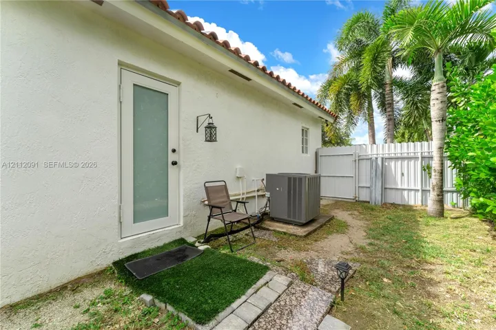 Exterior side yard featuring white stucco siding, a glass-panel door, terracotta roof tiles, a white vinyl privacy fence, and mature palm trees