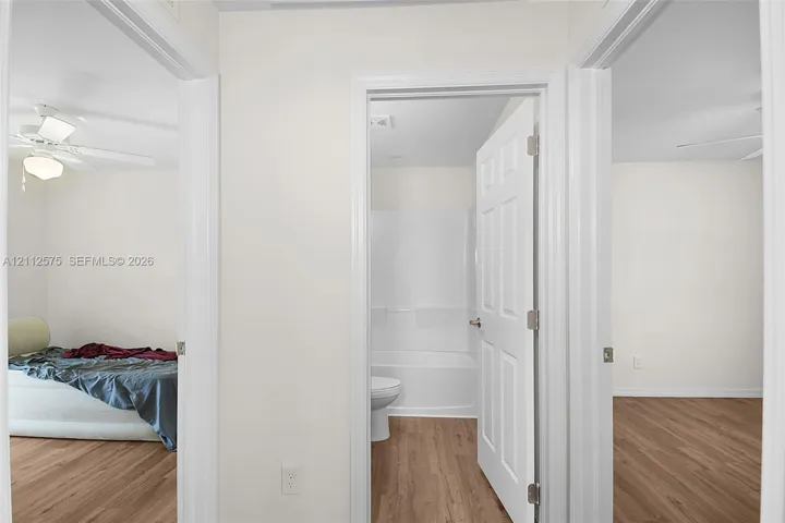 Interior hallway featuring wood-finish flooring, white six-panel doors, and a bathroom equipped with a white tub-shower surround