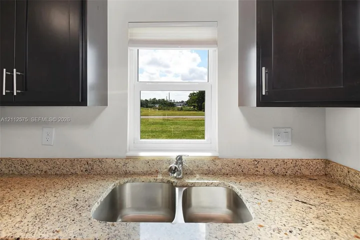 Deep-toned cabinetry featuring vertical pulls, speckled stone countertops, dual-basin stainless steel sink, and a centralized window with integrated shade