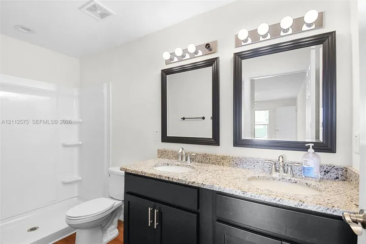 Dual-sink vanity with a stone-finish countertop, dark cabinetry, matching wall-mounted mirrors, and dual four-light fixtures, accompanied by a fiberglass shower surround